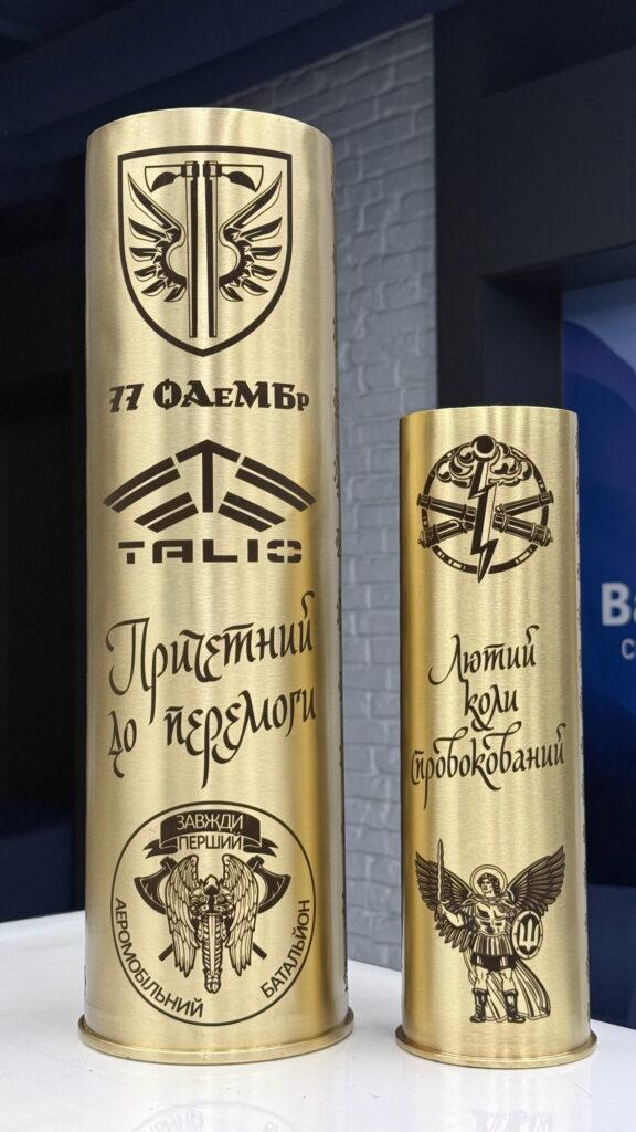 Латунна гільза з гравіюванням логотипу TALIS, шеврону 77 ОАеМБр та написом «Причетний до перемоги», поряд — мала гільза з Архангелом Михаїлом