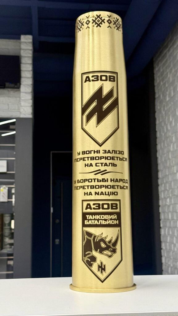 Гільза з орнаментом вишиванки на горлі, щитом АЗОВ, девізом «У вогні залізо перетворюється на сталь — у боротьбі народ перетворюється на націю» та емблемою танкового батальйону з носорогом. Символіка кількох рівнів на одній гільзі.