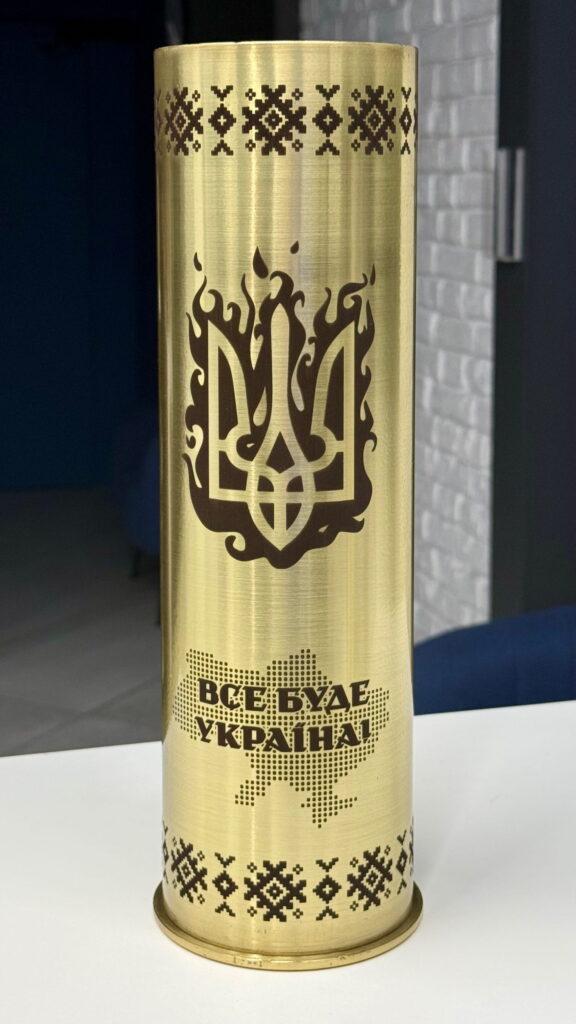 Тризуб у язиках полум'я — потужний і виразний образ. Пиксельна вишиванка зверху і знизу, точкова карта України з написом «Все буде Україна!». Гільза, яка говорить про силу та незламність.