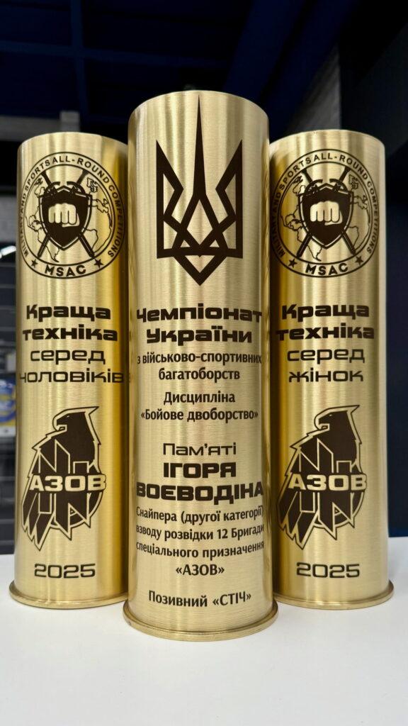 Три гільзи: центральна — меморіальна пам'яті Воєводіна, бокові — номінації «Краща техніка серед чоловіків» та «Краща техніка серед жінок» із щитом-орлом АЗОВ і роком «2025».