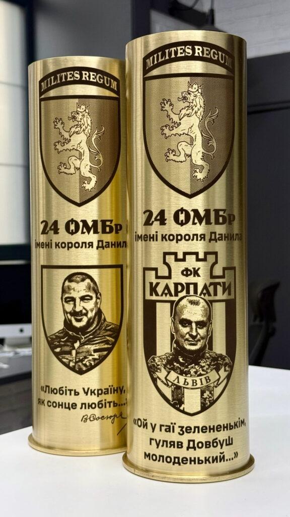 Дві артилерійські гільзи з шевроном 24 ОМБр імені короля Данила «Milites Regum», портрети командирів, шеврон ФК Карпати Львів, цитати Василя Симоненка та народна пісня про Довбуша Гільзи для двох командирів — однаковий стиль, різний зміст. На одній — цитата Симоненка «Любіть Україну, як сонце любіть», на іншій — народна «Ой у гаї зелененькім». Шеврон Карпат підкреслює регіональну ідентичність.