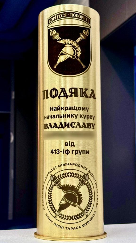 Артилерійська гільза з текстом «Подяка найкращому начальнику курсу від групи», шеврон Військового факультету міжнародних відносин та права ім. Тараса Шевченка ВІКНУ, зображення спартанського шолому Гільза як подяка викладачу від курсантів військового університету. Знизу — великий медальйон зі спартанським шоломом у лавровому вінку. Символіка класична і вічна.