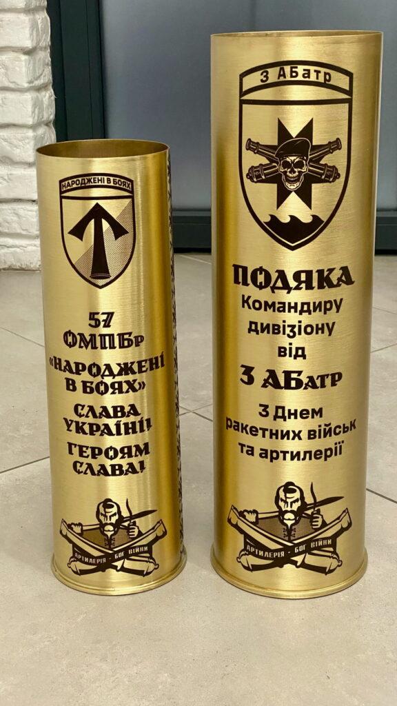 Дві артилерійські гільзи різного калібру поруч — менша гільза 57 ОМПБр «Народжені в боях» та велика гільза з подякою командиру дивізіону від 3 артилерійської батареї з Днем ракетних військ та артилерії, шеврон «Артилерія — бог війни» Наочна демонстрація різниці в калібрах. Більша гільза — подяка з Дня артилерії, шеврон із черепом та схрещеними гарматами і лозунгом «Артилерія — бог війни».