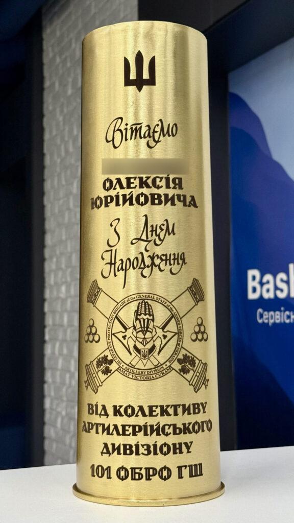 Артилерійська гільза зі стильним шрифтом для привітання Олексія Юрійовича з Днем народження, шеврон 101 бригади охорони Генерального штабу ЗСУ «Artillery Division. Amat Victoria Curam», тризуб угорі Поєднання каліграфічного декоративного шрифту для імені та класичного гарнітуру для підпису від колективу. Шеврон дивізіону з латинським девізом «Amat Victoria Curam» — «Перемога любить підготовку».